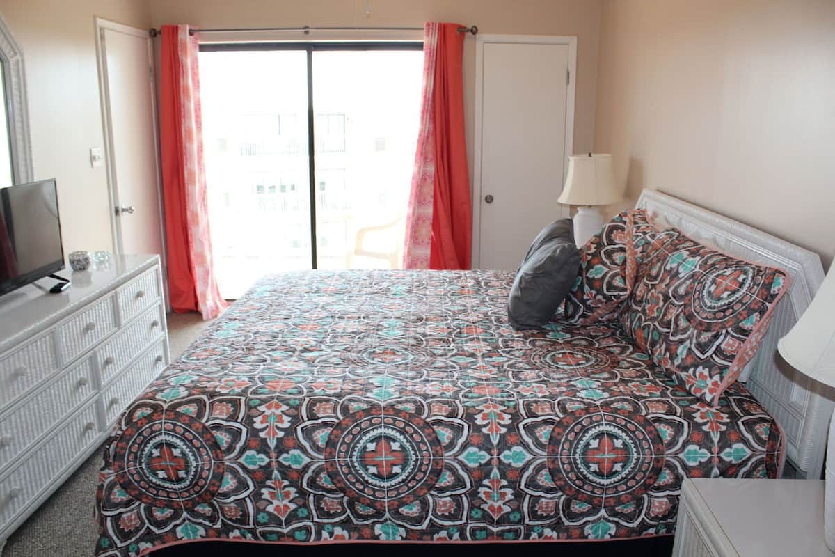 A bedroom with a bed, a dresser, a TV, a lamp, and a view of the outside.