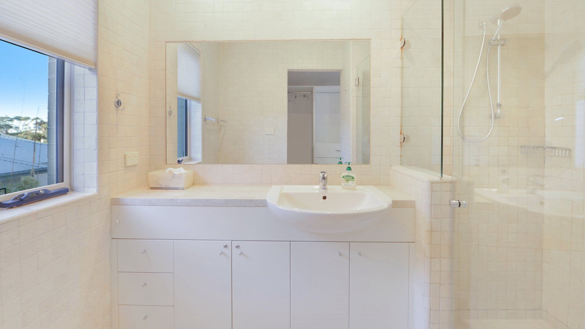 Un baño con lavamanos blanco, espejo y ducha. Hay una ventana a la izquierda y se ve un edificio desde el exterior.