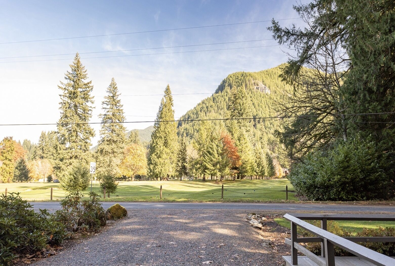 A gravel driveway leading to a wooden fence, with a mountain and dense forest in the background.