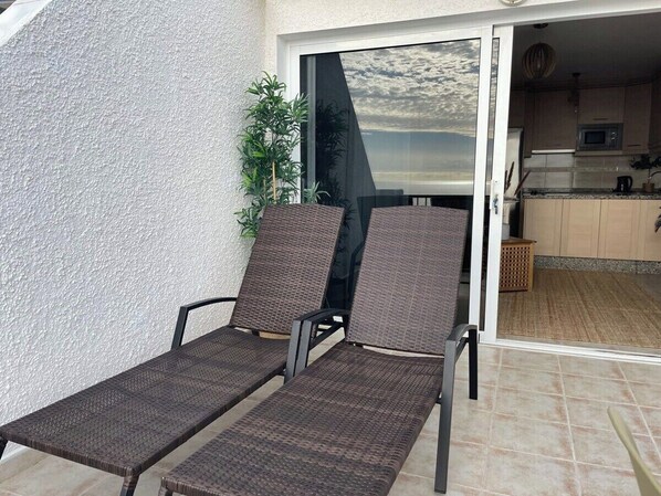 A balcony with two wicker lounge chairs, a potted plant, and a view of the kitchen and living area. - Ocean view apartment (Santiago del Teide)