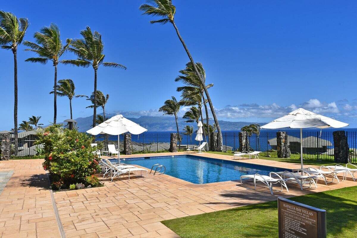 A pool area with lounge chairs and umbrellas, overlooking a body of water with palm trees.