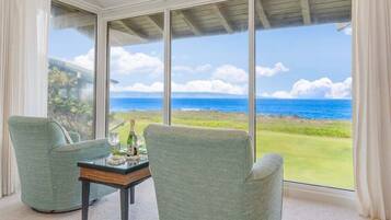 A room with a large window offering a view of the ocean and a grassy landscape. There are two chairs and a small table with a bottle and glasses.