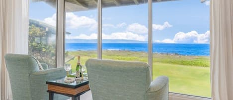 Et værelse med et stort vindue der viser udsigt over havet og et græslandskab. Der er to stole og et lille bord med en flaske og glas.