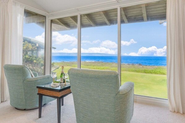 A room with a large window offering a view of the ocean and a grassy landscape. There are two chairs and a small table with a bottle and glasses.
