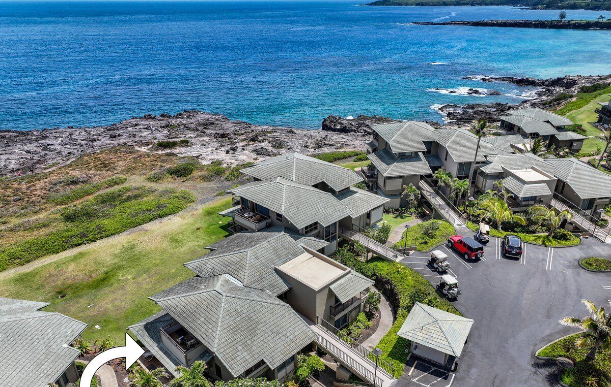 An aerial view of Kapalua Bay Villas 35G1, featuring a cluster of modern villas with sloped roofs, a parking area, and a view of the ocean.
