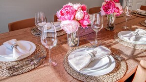 Een eettafel gedekt met witte borden, bestek en glazen wijnkelken, omringd door geweven tafelonderzetters en roze pioenrozen in glazen vazen.