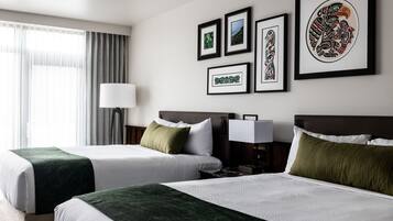 A hotel room with two beds, a large window with curtains, and several framed artworks on the wall.