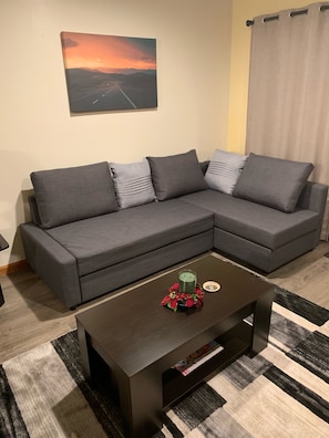 Ein Wohnzimmer mit einem dunkelbraunen Couchtisch, einem grauen Ecksofa mit vielen Kissen und einem Gemälde, das einen Sonnenuntergang zeigt.