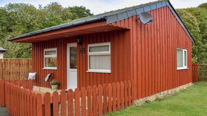 Ein rotes Holzhaus mit weißer Tür, zwei Fenstern, einer kleinen Veranda und einem eingezäunten Garten.
