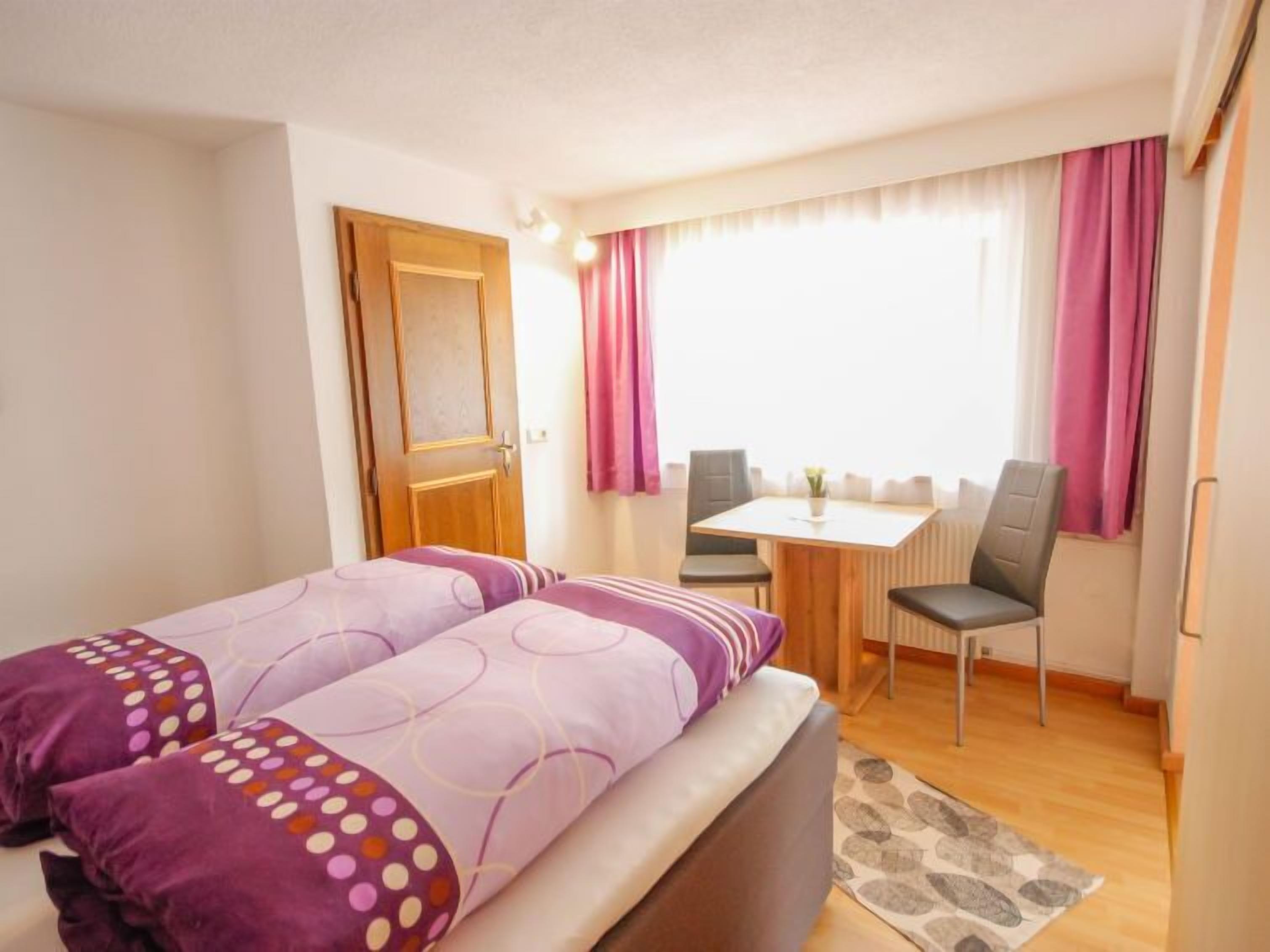 A bedroom with two beds, a wooden door, a small table with a chair, and a window with pink curtains.