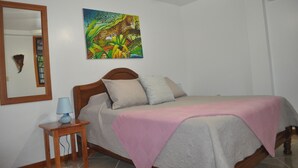 A bedroom with a bed, a bedside table with a lamp, a mirror, and a painting on the wall. - Latitude 17 Garden Room, heart of Hopkins Village, Belize (Hopkins Village)