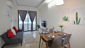 Ein modernes Wohnzimmer mit grauem Sofa, roten Kissen, einem Esstisch für zwei Personen und einem an der Wand montierten Fernseher.