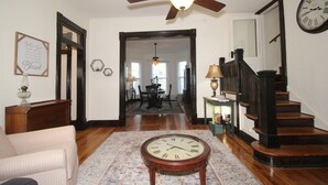 A spacious living room with hardwood floors, a ceiling fan, a large clock, and a staircase leading to another level. - Tastefully restored historic home in quaint town, 20 minute drive to Columbia MO (Centralia)