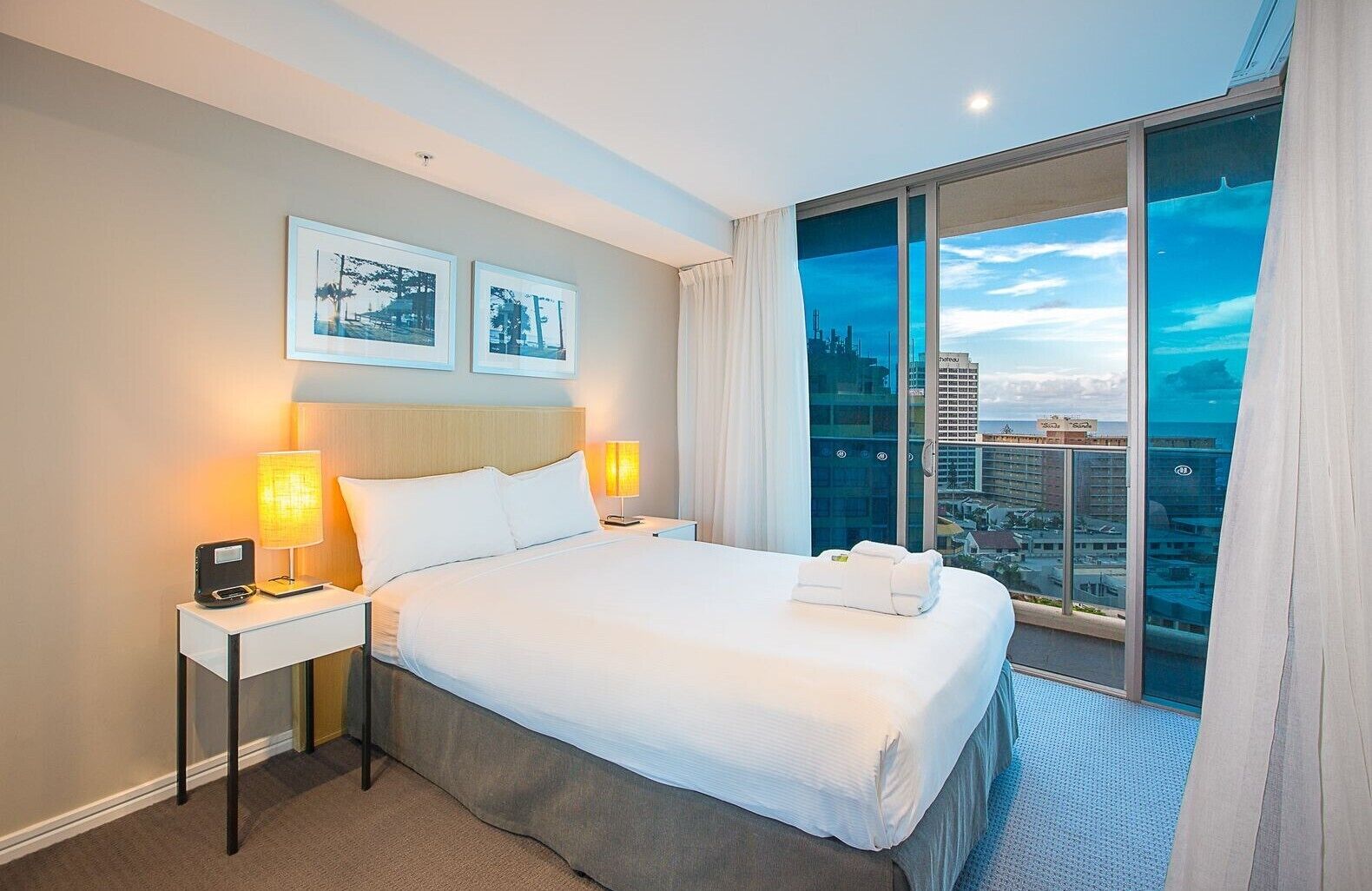 Habitación de hotel con una cama grande, dos lámparas de noche, una mesita de noche, vistas de la ciudad a través de ventanas de piso a techo y dos cuadros en la pared.