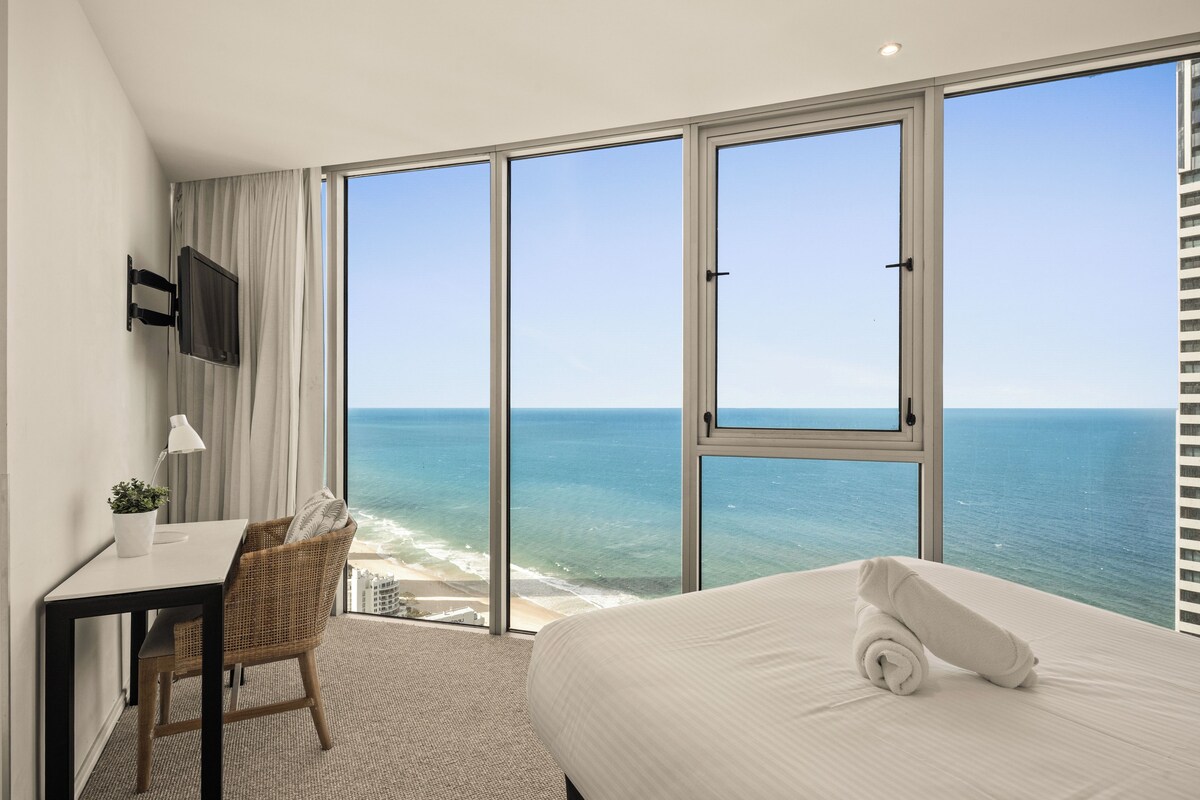 Habitación de hotel con un ventanal grande, una cama, un escritorio, una silla y un televisor.