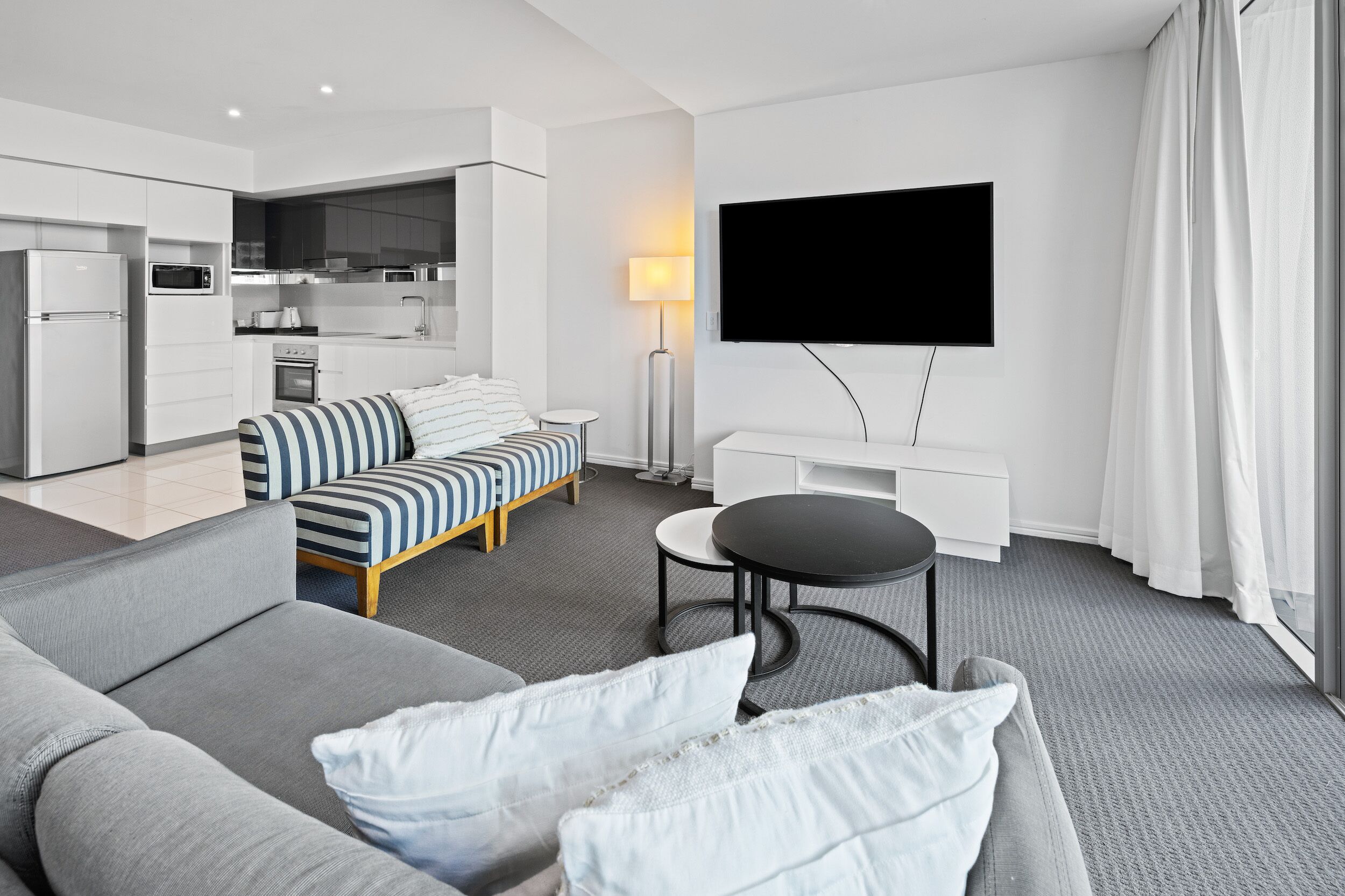 A modern hotel room with a grey sofa, a striped sofa, a small round table, a TV mounted on the wall, and a kitchen area in the background.