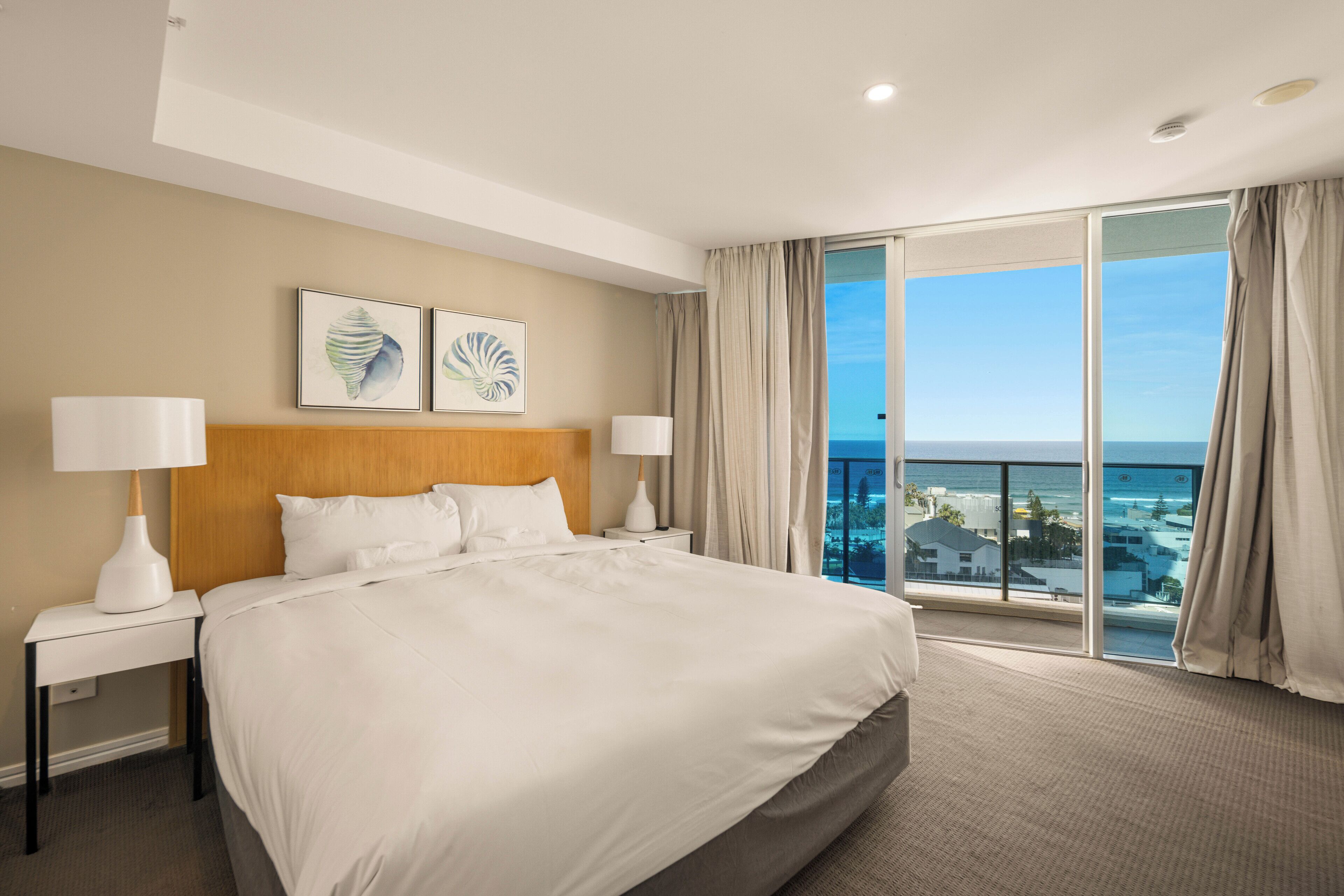 A hotel room with a large bed, two bedside tables with lamps, a wooden headboard, and a large window with a view of the ocean.