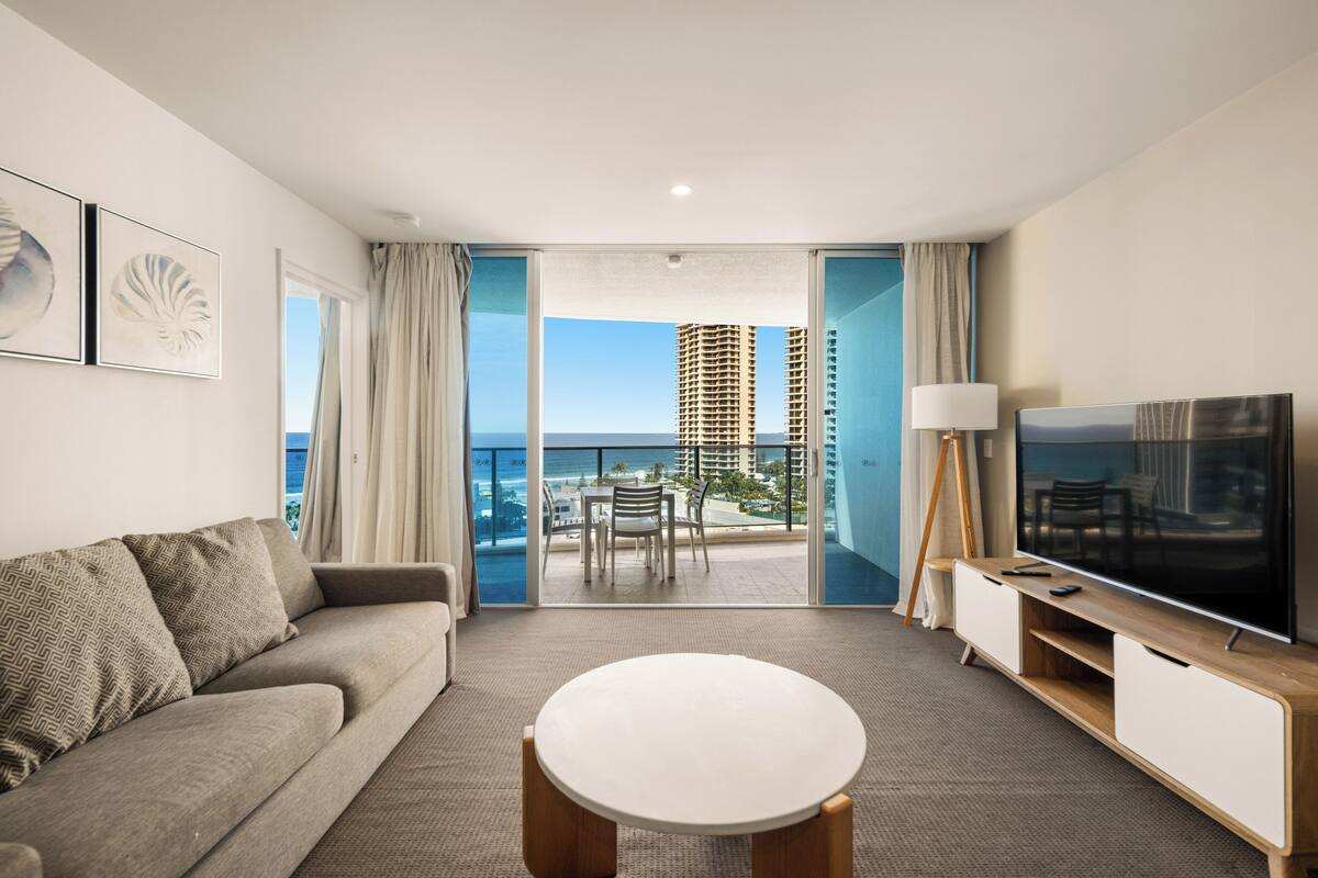 Una habitación de hotel moderna con balcón, sofá, televisor y mesa de comedor.