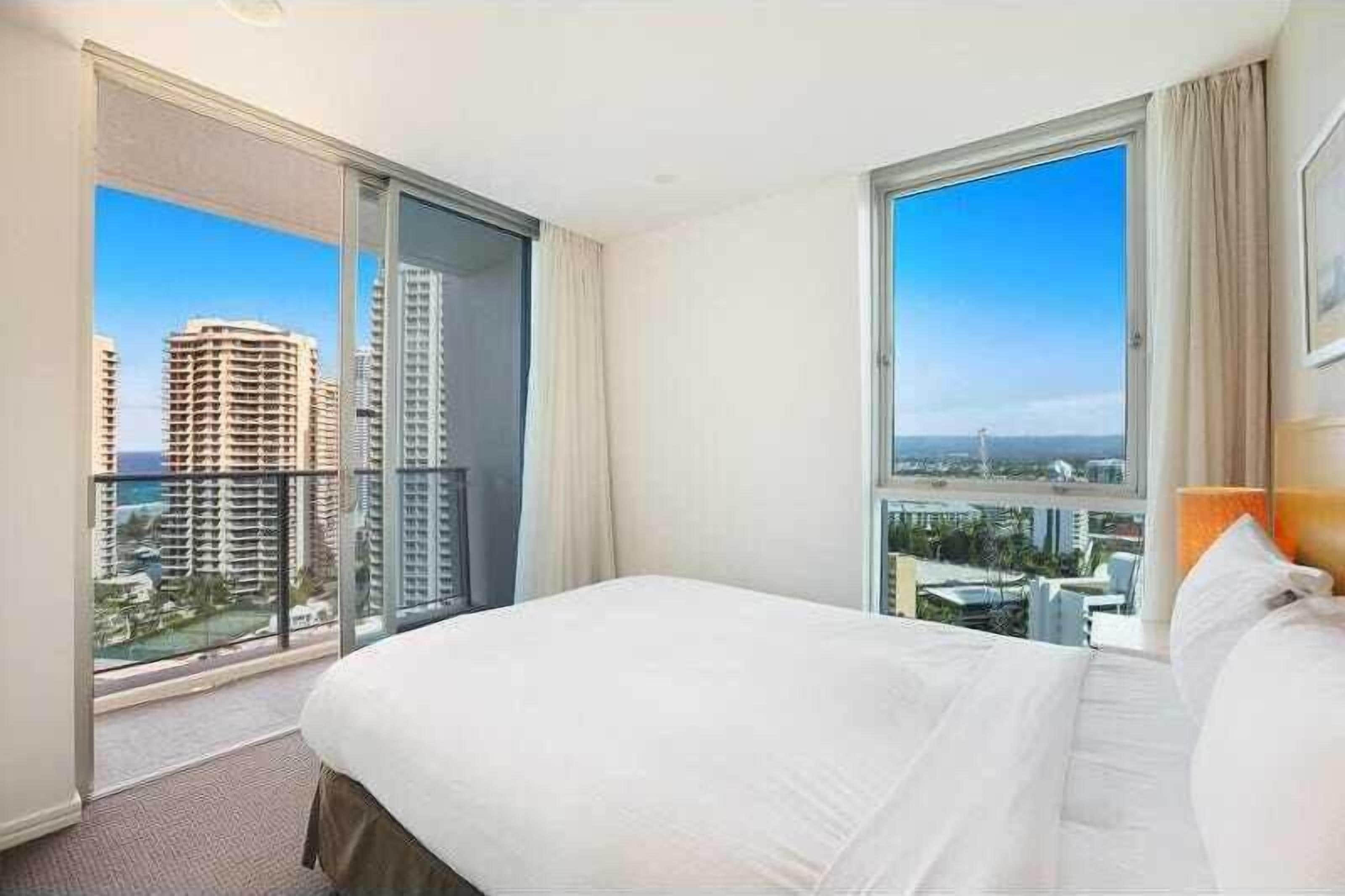 A hotel room with a large bed, a sliding glass door to a balcony, and a view of city buildings.