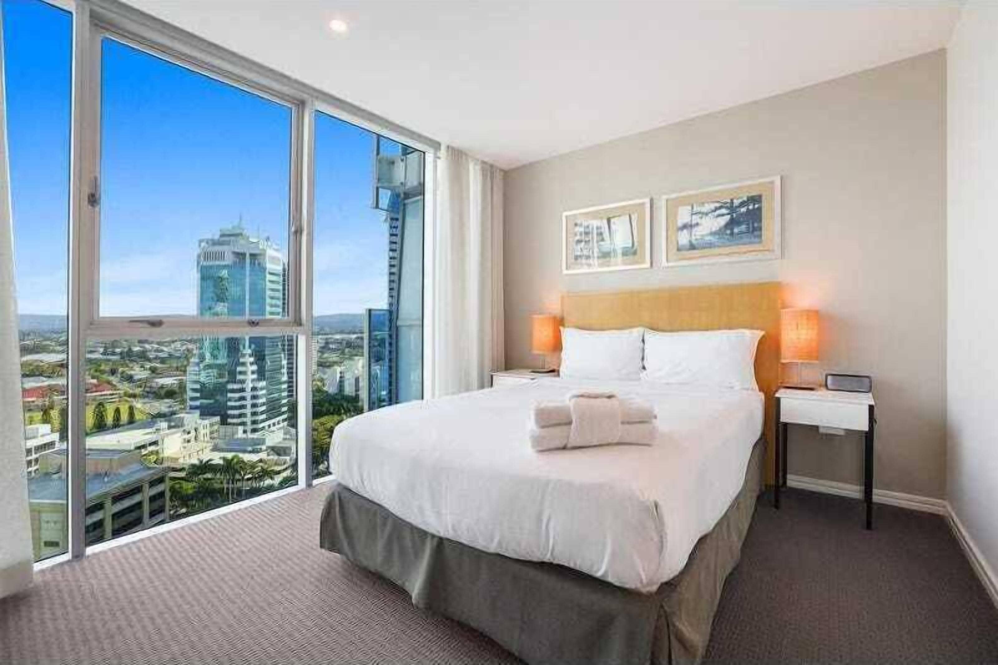 A hotel room with a large bed, two bedside tables, and a view of a cityscape through large windows.