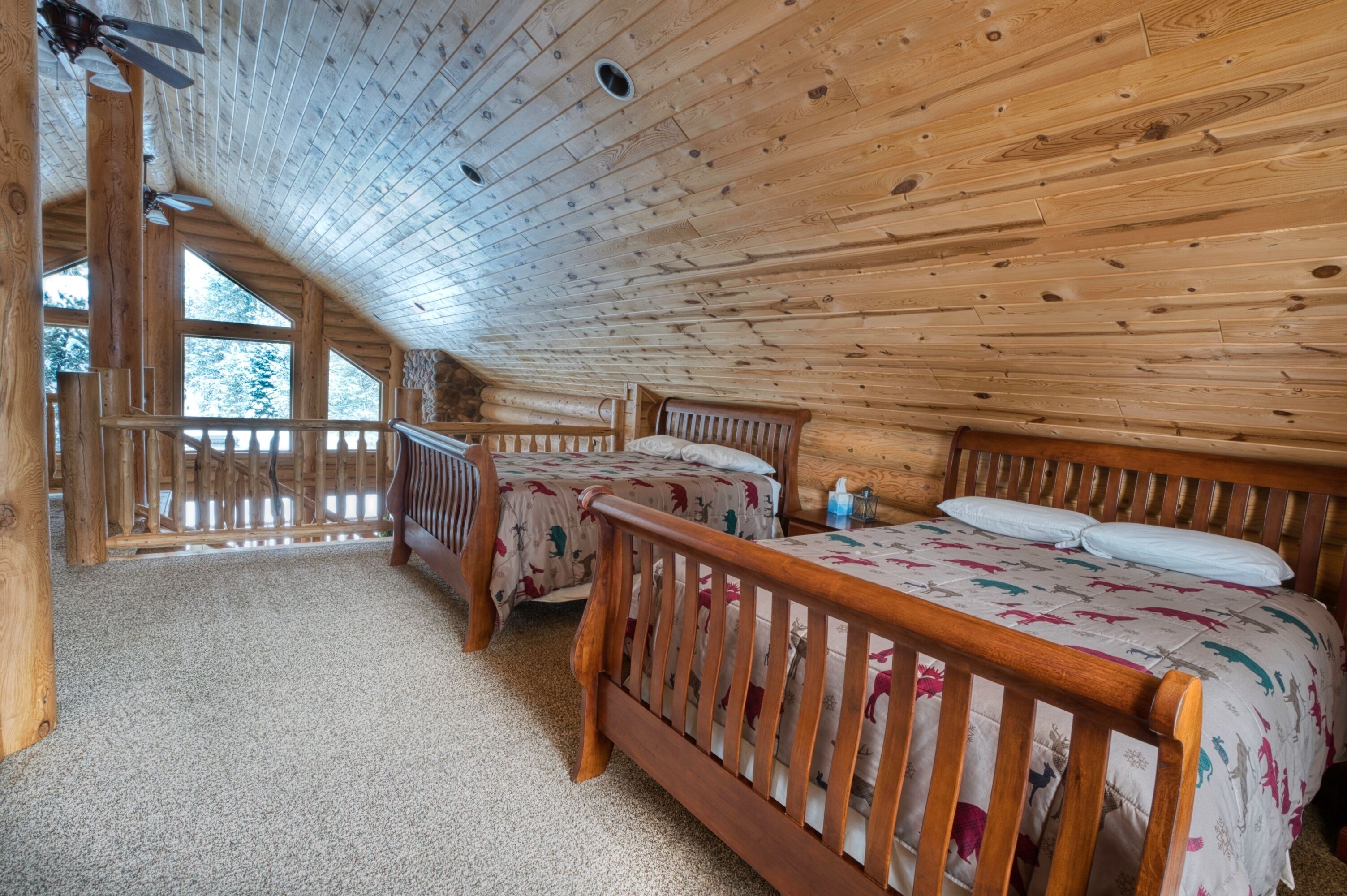 A cabin with two wooden beds, a ceiling fan, and a view of trees through large windows.