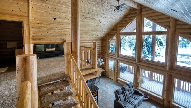 Interior de una cabaña de troncos con una escalera de madera, una sala de estar con sofá y ventanas amplias.