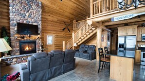 Un acogedor interior de cabaña de madera con chimenea de piedra, una zona de estar confortable, una cocina con electrodomésticos modernos y una escalera que lleva a un altillo.