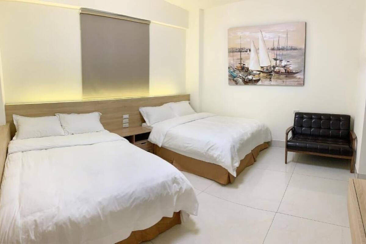 A hotel room with two beds, a bench, and a painting of sailboats on the wall.