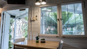 Una habitación con una mesa de madera, dos lámparas colgantes, un televisor montado en la pared y una ventana con vistas a zonas verdes.