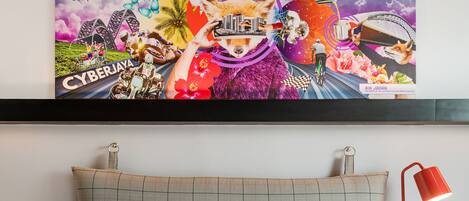 A hotel room with a large bed, two pillows, a red lamp, and a colorful artwork above the bed.