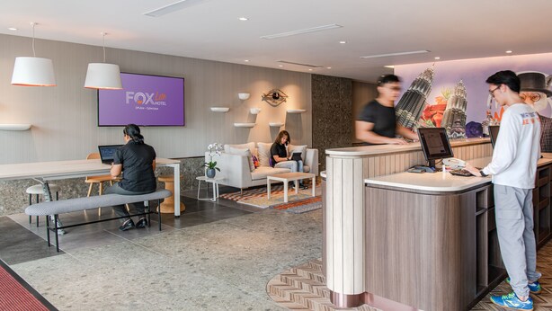 Een receptiehal van een hotel met een modern design, inclusief een receptiebalie, zithoek en een groot scherm met de naam van het hotel.