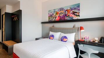 Habitación de hotel con cama, escritorio, silla, mesita de noche, lámpara y un cuadro en la pared.