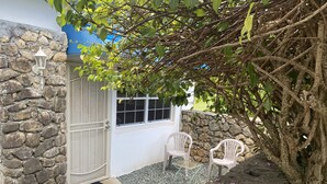 Une entrée avec un mur en pierre et une porte blanche, deux chaises en plastique blanches et un petit jardin agrémenté de gravier et d’un arbre.