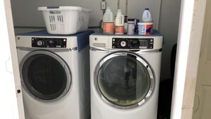 Deux machines à laver à chargement frontal se trouvent dans une buanderie, avec un panier et des bouteilles de lessive posés sur le dessus.