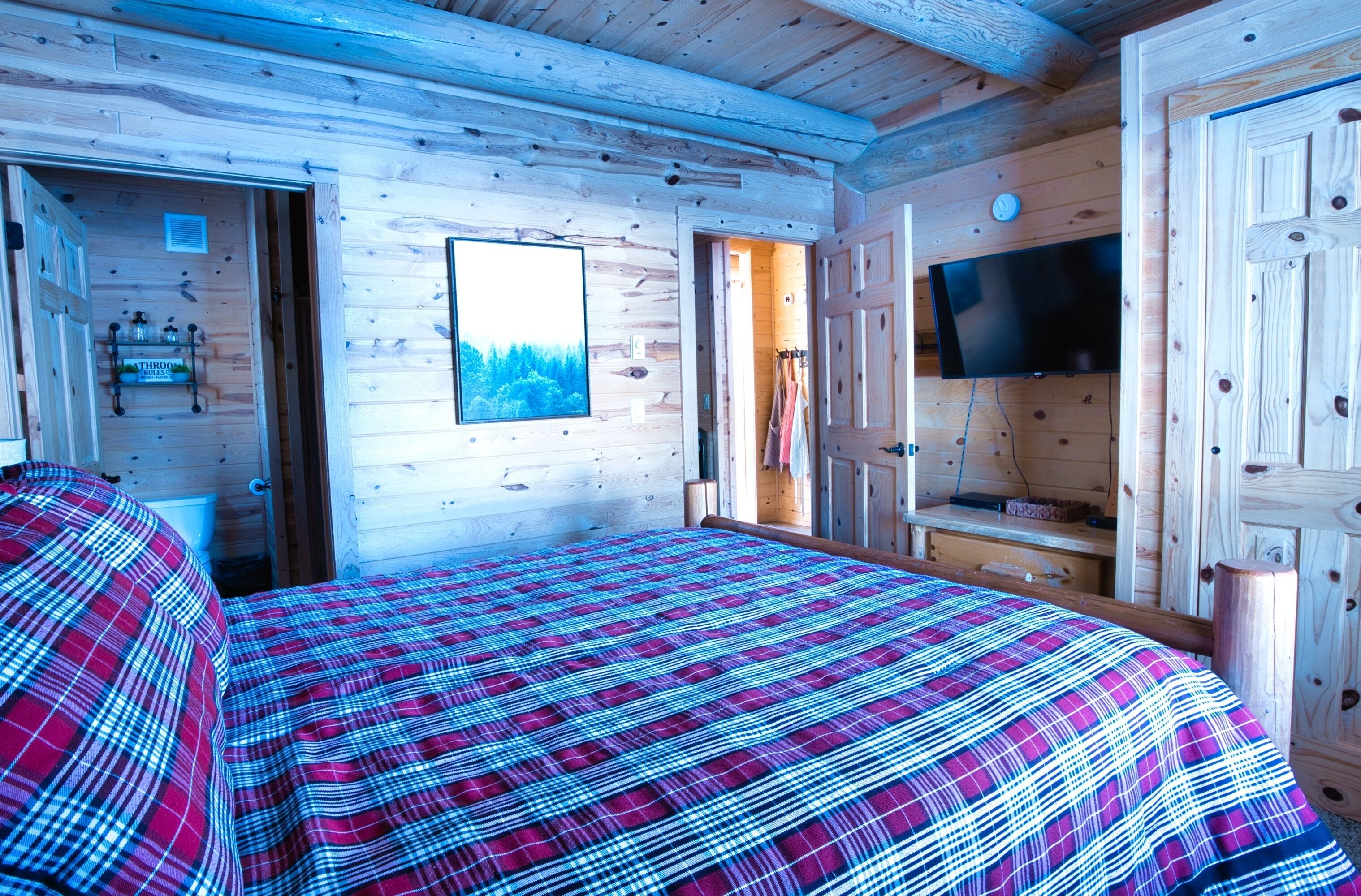 Une chambre à coucher dans un chalet en bois, avec une couverture à carreaux sur le lit, une télévision et une fenêtre donnant sur les arbres.