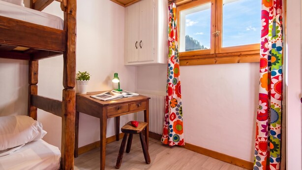 Een studentenkamer met een stapelbed, een bureau met lamp, een klein krukje, een radiator en een raam met gordijnen.