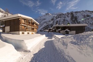 Un chalet de montagne enneigé, composé de plusieurs bâtiments, avec un ciel dégagé et une piste de ski en arrière-plan.