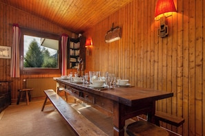 A wooden interior with a dining table set for six, a bench, a bookshelf, and a window with curtains.