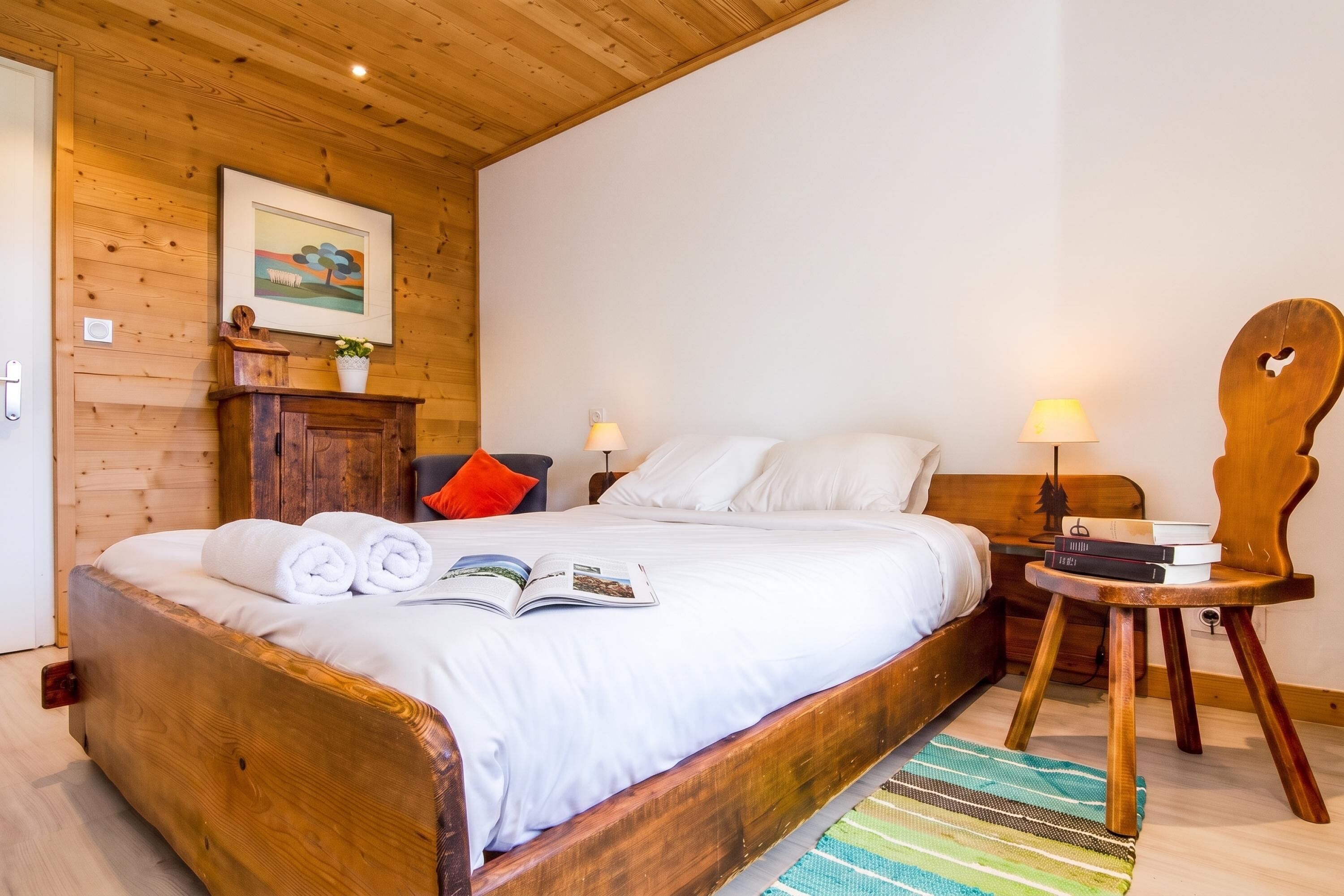 A wooden-framed bed with white bedding, a bedside table with a lamp, and a small wooden chair.