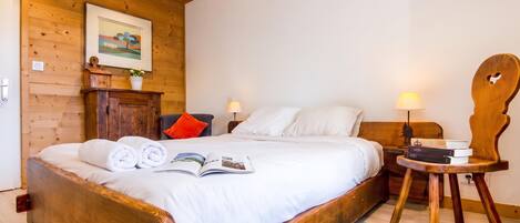 A wooden-framed bed with white bedding, a bedside table with a lamp, and a small wooden chair.