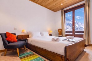 Een slaapkamer met een houten bed, een stoel, twee bedlampen, uitzicht op besneeuwde bergen door een raam, en een gestreept vloerkleed.