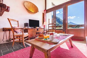Ein Zimmer mit einem Holztisch, gedeckt für das Frühstück, einem Fernseher und Blick auf Berge durch große Fenster.