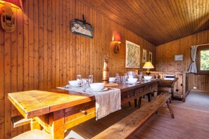 A wooden interior with a long dining table set for a meal, wooden walls, and a window with curtains.