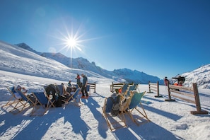 A snow-covered mountain area with people sitting on folding chairs, a wooden fence, and a clear blue sky.