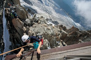 A person climbing a rocky mountain path with a rope, overlooking a glacier.