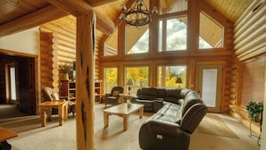 A log cabin interior with a large living area, wooden furniture, and a view of autumn trees through large windows.