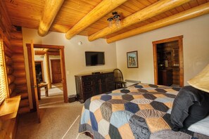 A log cabin bedroom with a wooden ceiling, a quilted bedspread, a dresser, a television, and a painting on the wall.