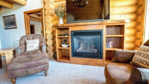 A cozy log cabin interior with a fireplace, leather armchairs, and a bookshelf.