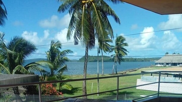 A terrace with a table covered by a patterned tablecloth, two wooden chairs, and a view of the sea and palm trees. - Gîte Océania (Wallis Island)