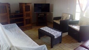 A living room with a sofa, a television, a wooden cabinet, and a wicker chair. - Gîte Océania (Wallis Island)